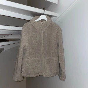 Beige teddyjacka med dragkedja - Mysig beige teddyjacka med dragkedja framtill. Jackan har långa ärmar och en hög krage för extra värme. Perfekt för kyliga dagar.