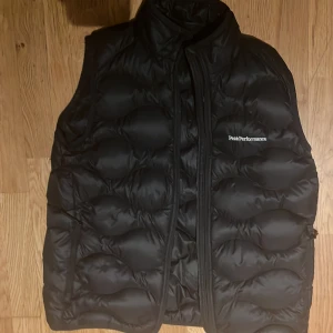 Svart dunväst från Peak Performance - Säljer en svart dunväst från Peak Performance. Västen har en dragkedja framtill och är perfekt för kyligare dagar. Den är stilren och lätt att matcha med andra plagg.Storlek 170 och den är knappt använd.Nypris är 1200 kr