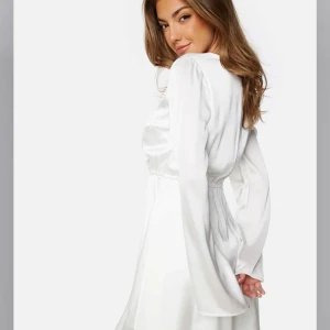 Vit omlottklänning  - Elegant vit omlottklänning  i satin med långa ärmar och v-ringning. Perfekt för en stilren look med en mjuk och glansig finish. Klänningen har en elastisk midja för en bekväm passform. Perfekt som studentklänning!! Köpt inför studenten 2024 men användes inte!