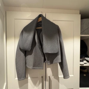 Grå vårjacka med halsduk - Snygg grå jacka från gina med en elegant och minimalistisk design. Jackan har en lös passform och är perfekt för höst och vår. Den har långa ärmar och en stilren look som passar till många olika outfits. Halsduk medförs