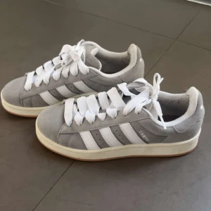 Grå Adidas Campus 00 sneakers  - Snygga grå Adidas Campus sneakers med vita ränder och breda snören. Skorna har en klassisk design med en bekväm sula i gummi. Perfekta för en stilren och avslappnad look. Skorna är andvnända vid 2 tillfällen 