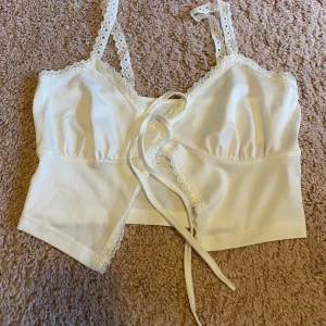 Vit spetsprydd topp - Säljer en söt vit korsettopp med spetsdetaljer och knytning framtill. Toppen har smala axelband och en croppad passform, perfekt för sommarens varma dagar. 