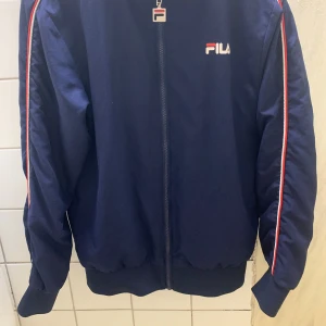 Blå träningsjacka från FILA - Säljer en snygg blå träningsjacka från FILA med dragkedja och ränder i rött och vitt längs ärmarna. Perfekt för sportiga aktiviteter eller en avslappnad stil. Jackan har en klassisk design med ribbade muddar och krage.absolut finns ingen skador 💝