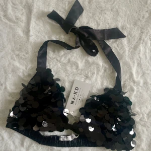 Svart paljett triangeltopp från NA-KD - Snygg svart triangeltopp från NA-KD täckt med glittrande paljetter. Den har knytband runt nacken och resårband på ryggen för justerbar bra passform. Perfekt under kavaj eller liknande. Aldrig använd därav prislapp kvar. Nypris 399