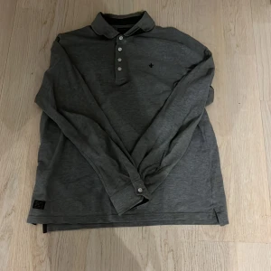 Grå rugbytröja  från Morris - Säljer en stilren grå långärmad pikétröja från Morris. Tröjan har en klassisk krage med knappar och en diskret broderad detalj på bröstet. Perfekt för en avslappnad men ändå uppklädd look.
