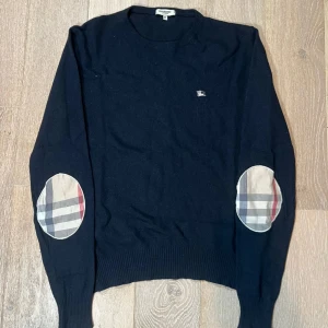 Svart tröja med rutiga armbågslappar - Snygg svart tröja från Burberry med klassiska rutiga armbågslappar. Tröjan har långa ärmar och en liten logga på bröstet. Perfekt för en stilren look. Skicka 8/10 Inga skador passar även M (tar även byten)