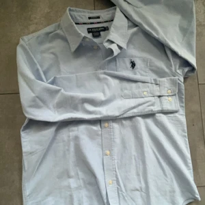 Ljusblå skjorta från U.S. Polo Assn. - Säljer en ljusblå skjorta från U.S. Polo Assn. med broderad logga på bröstet. Skjortan har långa ärmar och knappar framtill. Den är tillverkad i en bomullsblandning med stretch för extra komfort.