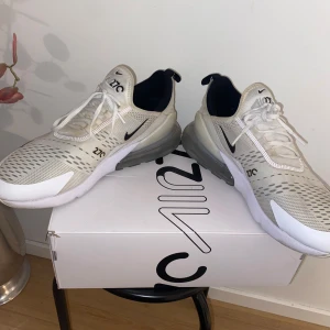 Nike Air Max 270  - Snygga Nike Air Max 270 sneakers i vit med svarta detaljer och vit sula. Skorna har en luftig ovandel med snörning och den ikoniska Air Max-dämpningen i hälen för extra komfort.