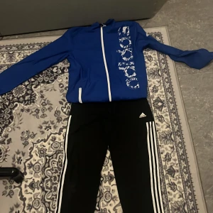 Blå träningsjacka från Adidas - Säljer en blå träningsjacka från Adidas med vit dragkedja och stort tryck på framsidan. Jackan har långa ärmar och är perfekt för sportiga aktiviteter. Den är både snygg och funktionell för träning. Priset kan diskuteras 