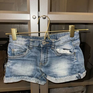 Blå low waist  jeansshorts med slitningar - Snygga low waist blå jeansshorts med slitningar och fransiga kanter. De har en klassisk femficksdesign och knappgylf. Perfekta för en avslappnad stil.