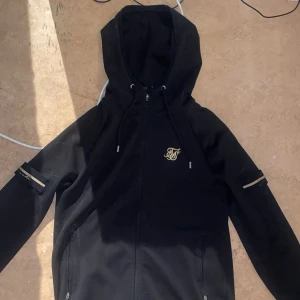 Svart hoodie från SikSilk - Snygg svart hoodie från SikSilk med dragkedja och broderad logga på bröstet. Den har långa ärmar med dekorativa band och en justerbar huva. Perfekt för en casual look.