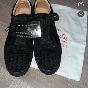  Christian Louboutin skor - Snygga svarta sneakers från Christian Louboutin med ikoniska röda sulor. Skorna har en unik design med svarta nitar på ovandelen och snörning framtill. Perfekta för att ge en edgy touch till din stil.