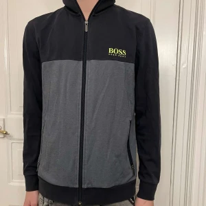 Svart och grå tröja från Boss - Säljer en snygg svart och grå tröja från Boss i mycket bra skick. Den har en dragkedja framtill och två praktiska fickor med dragkedjor. Perfekt för höst och vår! Stor logga på bröstet.