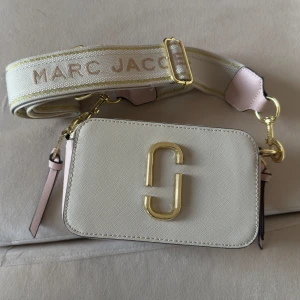 Axelväska från Marc Jacobs - Snygg beige axelväska från Marc Jacobs med ett stort guldigt 'J' på framsidan. Väskan har en bred axelrem med märkesnamnet i guld och rosa detaljer. Perfekt för att ge en lyxig touch till din outfit! Som ny! 