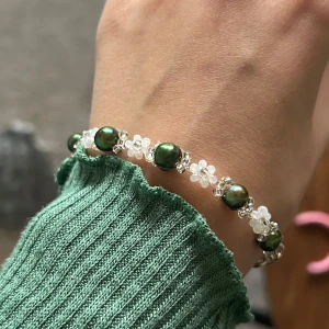 Armband med gröna pärlor och blommor - Snyggt handgjort armband med gröna pärlor och små vita blommor. Perfekt för att ge en touch av elegans till vilken outfit som helst. Armbandet har en justerbar kedja för perfekt passform. Passar både till vardags och festliga tillfällen. Gjort i äkta rocaillespärlor (glas) och kedjan är försilvrad. 