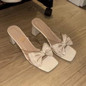 Beige klackskor med rosett - Snygga beige klackskor med en stor rosett framtill. De har en fyrkantig tå och transparenta remmar som ger en modern touch. Perfekta för festliga tillfällen eller en utekväll. Klacken är stabil och bekväm att gå i. Storlek 39 men funkar för 38