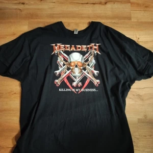 Svart Megadeth t-shirt - Säljer en svart Megadeth t-shirt med tryck på både fram- och baksidan. Framsidan har ett coolt dödskallemotiv med texten 'Killing is my business...'. Baksidan listar låttitlar från bandet. Perfekt för rockälskare! T-shirten är kortärmad och i bra skick.