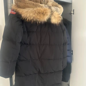 PJS jacka longbear  - Säljer en svart dunjacka från Parajumpers i mycket bra skick. Jackan har en mysig pälsdetalj på huvan och en praktisk dragkedja framtill. Perfekt för kalla vinterdagar. Den har också en cool patch på ärmen som ger en extra touch av stil.