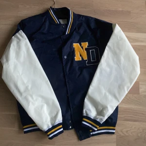 Blå och vit bomberjacka med ND-broderi - Säljer en snygg blå och vit bomberjacka med broderat ND-märke på bröstet. Jackan har långa vita ärmar och blå kropp, med ribbstickade muddar i blått, vitt och gult. Perfekt för höst och vår!