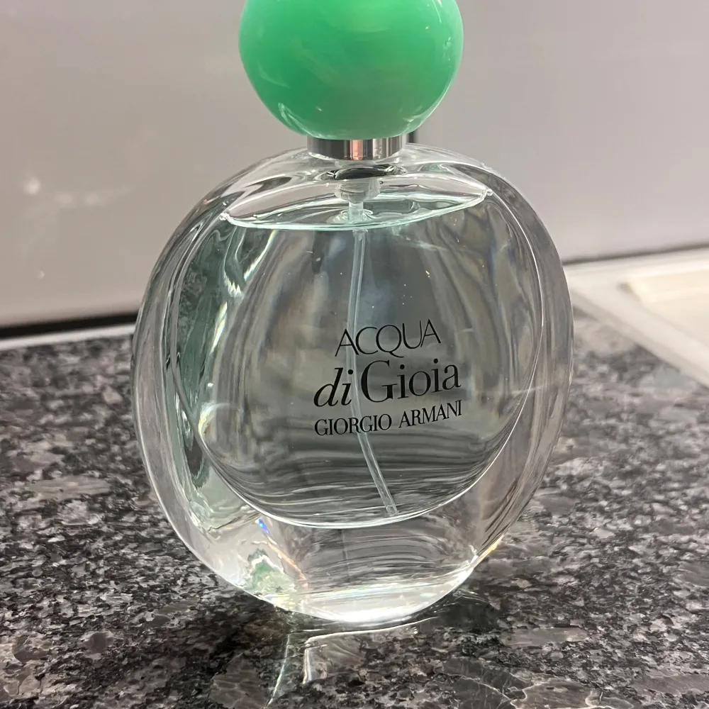 Säljer en elegant och fräsch parfymflaska av Acqua di Gioia från Giorgio Armani. Flaskan är genomskinlig med en rund form och har en stilren, mintgrön kork. Perfekt för dig som älskar en fräsch och naturlig doft. Passar både till vardags och speciella tillfällen.. Parfym.