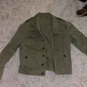 Militär grön fieldjacket  - Säljer en snygg olivgrön jacka från Mango i storlek M. Jackan har en klassisk design med knappar framtill och flera praktiska fickor. Perfekt för höst och vår, och ger en stilren look. Passar både till vardags och lite mer uppklädda tillfällen. Jackan är använd 1 gång och är i super fint skick. Priset är ej hugget i sten, skriv ifall de finns frågor