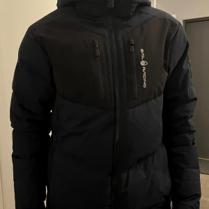 Svart pufferjacka från Sail Racing - Säljer en svart pufferjacka från Sail Racing i bra skick. Jackan har en dragkedja framtill och två bröstfickor med dragkedjor. Perfekt för kalla vinterdagar med sin varma och vadderade design. Huvan är justerbar och jackan har en stilren look. Det finns ett par små defekter på jackan