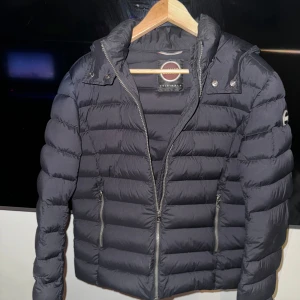 Colmar dunjacka - Säljer en snygg svart pufferjacka från Colmar som är i bra skick. Perfekt för höst och vinter med sin varma och quiltade design. Den har en huva för extra skydd mot kyla. Passar perfekt för vardags användning. Har fixats hos skräddare som syns i bild 3 och 4, inget som syns om man inte tittar riktigt noga (lågt pris på grund av defekterna)