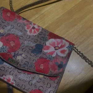 Blommig axelväska från Guess - Säljer en superfin axelväska från Guess med blommigt mönster. Väskan har en beige bas med röda och rosa blommor samt Guess-logga i bakgrunden. Den har en kedjerem och är perfekt för både vardag och fest. Väskan är i mycket bra skick och redo för nya äventyr! 🌸 kan mötas upp i Stockholm 