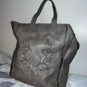 Grå axelväska med glitter - Säljer en snygg grå axelväska i skinn med gulliga glitter pluppar i form av vingar på framsidan. Väskan har dragkedjor på sidorna och ett rymligt inre med flera fack. Perfekt för både vardag och fest! Väl använd 