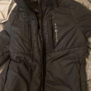 Svart pufferjacka från Sail Racing - Säljer en svart pufferjacka från Sail Racing i bra skick. Jackan har en dragkedja framtill och två praktiska bröstfickor med dragkedjor. Perfekt för kyliga dagar och har en stilren design som passar till det mesta. Hör av dig vid intresse!