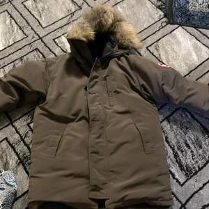 Säljer en brun parkajacka från Canada Goose i mycket bra skick. Jackan har en varm pälsfodrad huva och dragkedja med knappar. Perfekt för kalla vinterdagar. Den är långärmad och quiltad för extra värme. Passar både höst och vinter.