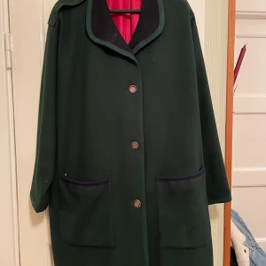 Stilig höstkappa - Välvårdad mörkgrön 80-talskappa med snyggt fall i 80% ull. Rött foder, dekorativa knappar och knappslå över sprund på vardera sidan. Raglanärm. Svensktillverkad. Kemtvätt. 107 cm från krage till nederkant. Rymlig för storleken. 
