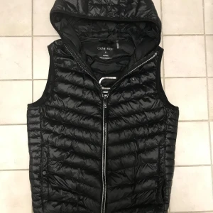 Svart dunväst Calvin Klein - Säljer en snygg svart dunväst från Calvin Klein i storlek S. Västen har en praktisk dragkedja och en huva, perfekt för kyliga dagar. Den är lätt, vilket gör den idealisk för att packa. Har inga defekter och är tvär snygg, återkom med frågor om ni har intresse😊