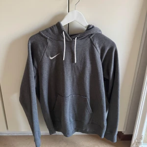 Grå hoodie från Nike - Säljer en snygg och bekväm grå hoodie från Nike. Den har en klassisk design med en vit Nike-logga på bröstet och en praktisk magficka. Perfekt för både vardag och träning. Huvan har justerbara snören för bästa passform. Passar perfekt till höst och vår!