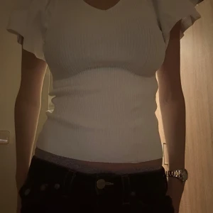 Vit ribbad topp - Säljer en superfin vit ribbad topp med korta ärmar och v-ringning. Toppen har en snygg passform som är tight och framhäver figuren. Perfekt för både vardag och fest! Den har en öppning i ryggen med snörning. Passar perfekt till jeans eller kjol.
