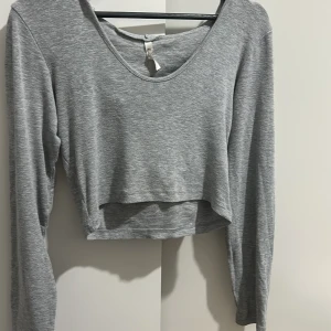 Grå croppad tröja från Kikiriki - Säljer en snygg grå croppad tröja från Kikiriki i ribbat material. Tröjan har långa ärmar och en djup rund halsringning. Perfekt för en avslappnad stil eller att matcha med högmidjade jeans. Passar bra till både vår och höst!