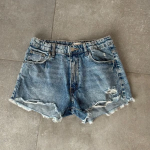 Blå jeansshorts från Gina Tricot - Snygga blå jeansshorts från Gina Tricot med slitna detaljer och fransiga kanter. De har en klassisk femficksdesign och knappgylf. Perfekta för en avslappnad stil.