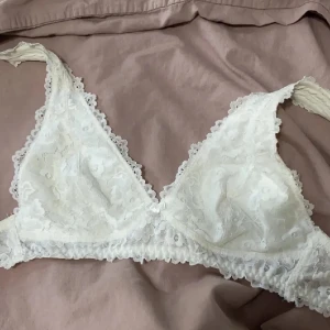 Vit spets bralette - Säljer en vacker vit spets bralette med triangelformade kupor och justerbara axelband. Den har en mjuk och romantisk design med hakknäppning bak. Perfekt för en bekväm och stilren look. Använd några gånger, nytvättad. Antingen från Lindex eller Kappahl
