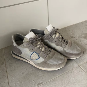 Grå sneakers från Philippe Model - Snygga grå sneakers från Philippe Model med en stilren design. Skorna har en kombination av mocka och syntetmaterial, med snörning och en diskret logga på sidan. Perfekta för en avslappnad stil. Skorna har tappat en del av sulan på ena skon (se bild), detta syns dock knappt när man har på dom. Skick 7/10, dom är ganska slitna, därav det väldigt låga priset. Perfekt för sommaren!