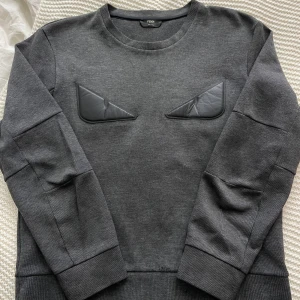 Grå tröja från Fendi ”Bug eye” - Säljer en stilren grå tröja från Fendi med unika detaljer på bröstet. Tröjan har långa ärmar och en rund halsringning. Perfekt för dig som vill ha något exklusivt i garderoben. Tillverkad i Italien. Nypris 9000kr.