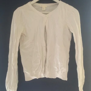 Vit kofta från H&M - Säljer en klassisk vit kofta från H&M i storlek XS. Koftan är tillverkad i 100% bomull och har långa ärmar med knappar framtill. Perfekt för lager-på-lager-stil eller som ett lättare ytterplagg.