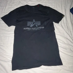 Svart t-shirt från Alpha Industries - Säljer en svart t-shirt från Alpha Industries med tryck på framsidan. T-shirten har en klassisk rund hals och korta ärmar. Perfekt för en avslappnad stil.