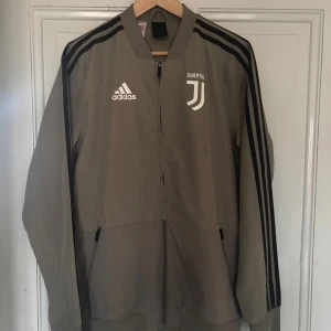 Juventus Träningsjacka - Säljer en träningsjacka från Adidas med Juventus-logga på bröstet. Jackan har svarta ränder längs ärmarna och en dragkedja framtill. Perfekt för träningar eller för vanlig daglig användning💪🏾💯 har en väldigt liten fläck på ena bröstet, men knappt synligt när man väl har på💯