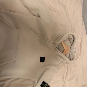 Beige hoodie från Stone Island - Säljer en snygg beige hoodie från Stone Island med deras ikoniska märke på ärmen. Hoodien har en känguruficka och justerbar huva med dragsko. Perfekt för en stilren och avslappnad look. Använd 1 gång då den är för liten för mig 