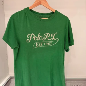 Grön t-shirt från Polo Ralph Lauren - Säljer en grön t-shirt från Polo Ralph Lauren i storlek M med tryck 'Polo RL Est 1967' på framsidan. Har ett litet hål under armen annars skulle jag säga att skicket är 8/10