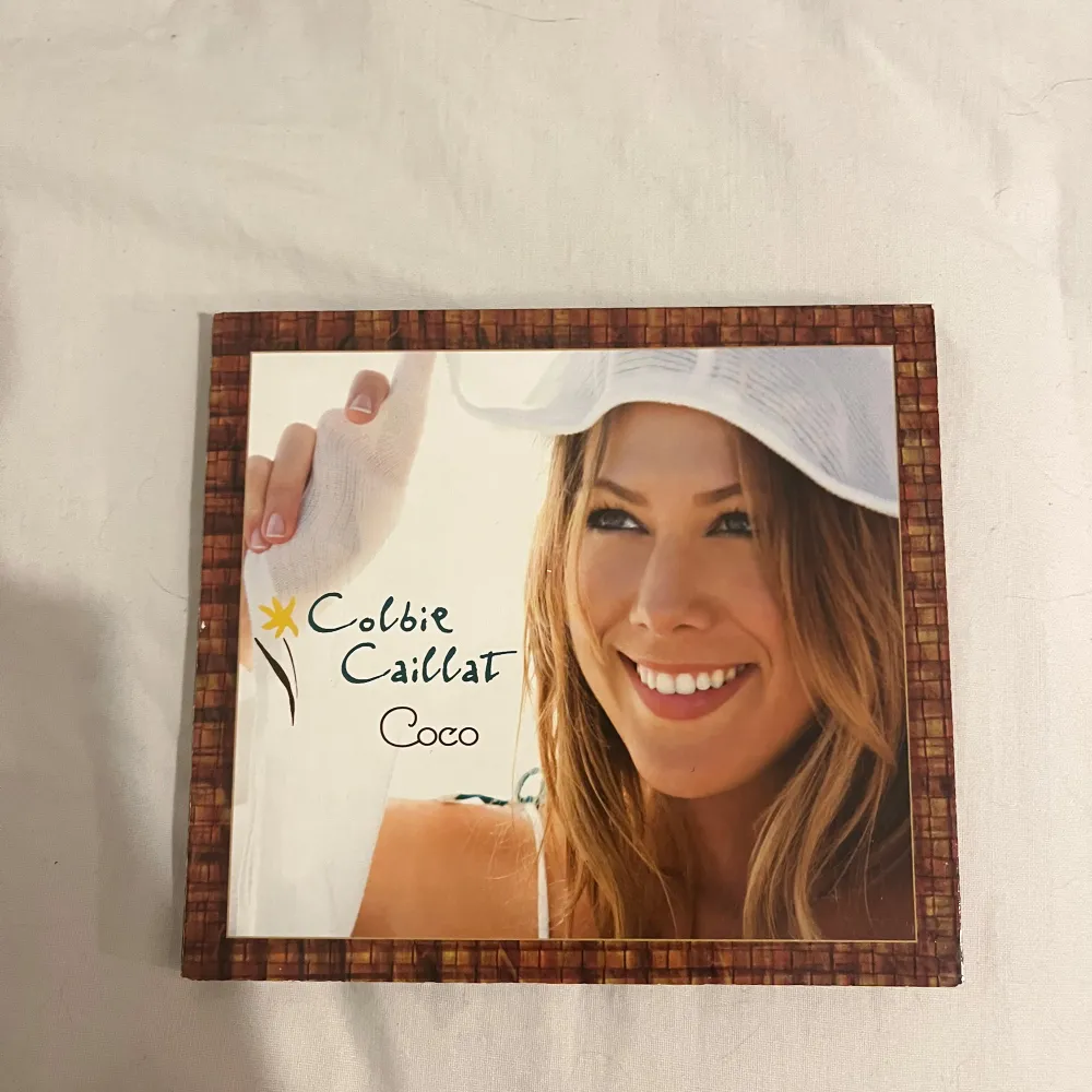 CD-albumet 'Coco' av Colbie Caillat. Omslaget har en bild av en leende kvinna med en vit hatt. Inuti finns bilder av artisten i olika miljöer samt låttexter. Skivan har en träliknande design med artistens namn och albumtitel tryckt i vitt.. Muu.