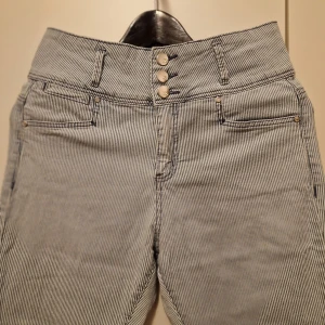 Randiga högmidjade jeans - Snygga högmidjade jeans med smala ränder i blått och vitt. De har en knappgylf med fyra knappar och dekorativa knappar vid bensluten. Perfekta för en stilren look.