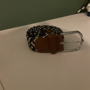 Svart, grått och vitt flätat bälte - Snyggt flätat bälte i svart, grå och vitt med bruna läderdetaljer. Bältet har ett silverfärgat spänne och passar perfekt för att ge en stilren touch till din outfit.