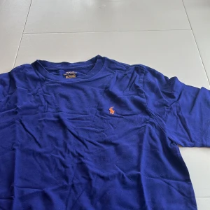 Blå t-shirt från Polo Ralph Lauren - Säljer en snygg blå t-shirt från Polo Ralph Lauren med det klassiska broderade logotypen i orange på bröstet. Perfekt för en avslappnad stil. T-shirten har korta ärmar och en rund halsringning.
