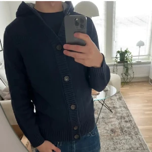 Stickad cardigan - Säljer en mörkblå stickad hoodie med knappar framtill. Tröjan har en mysig huva med ljusgrått foder och ribbade muddar vid ärmslut och nederkant. Storlek är M.